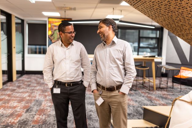 Two staff members stand chatting and laughing in our National Office with meeting rooms and tables in the background